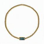 A December Birthstone Aqua Cubic Zirconia Baguette Bracelet by Skōsh Jewelry, featuring a strand of polished gold beads on a stretch cord with a rectangular blue baguette stone at the center, displayed against a white background.