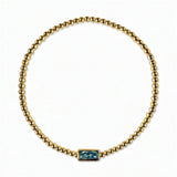 A December Birthstone Aqua Cubic Zirconia Baguette Bracelet by Skōsh Jewelry, featuring a strand of polished gold beads on a stretch cord with a rectangular blue baguette stone at the center, displayed against a white background.