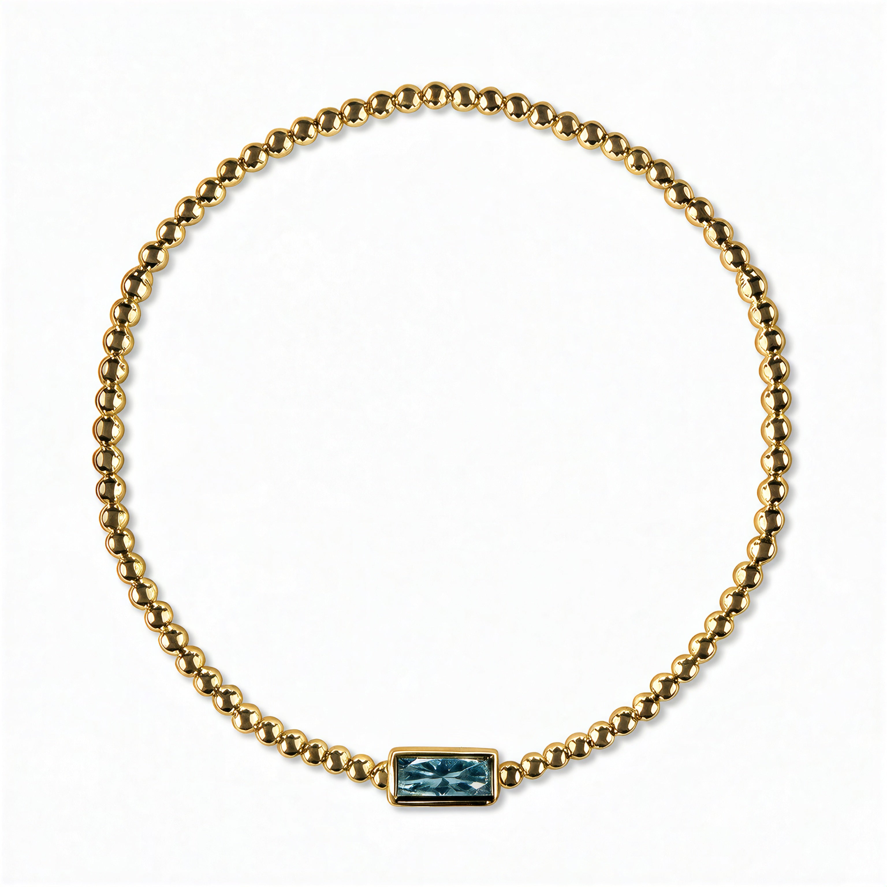 A December Birthstone Aqua Cubic Zirconia Baguette Bracelet by Skōsh Jewelry, featuring a strand of polished gold beads on a stretch cord with a rectangular blue baguette stone at the center, displayed against a white background.