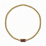 A February Birthstone Amethyst Cubic Zirconia Baguette Bracelet by Skōsh Jewelry, featuring a strand of polished gold beads on a stretch cord with a rectangular purple baguette stone at the center, displayed against a white background.