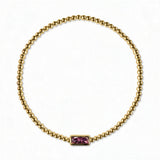 A February Birthstone Amethyst Cubic Zirconia Baguette Bracelet by Skōsh Jewelry, featuring a strand of polished gold beads on a stretch cord with a rectangular purple baguette stone at the center, displayed against a white background.