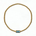 A March Birthstone Light Sapphire Cubic Zirconia Baguette Bracelet by Skōsh Jewelry, featuring a strand of polished gold beads on a stretch cord with a rectangular light blue baguette stone at the center, displayed against a white background.