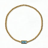 A March Birthstone Light Sapphire Cubic Zirconia Baguette Bracelet by Skōsh Jewelry, featuring a strand of polished gold beads on a stretch cord with a rectangular light blue baguette stone at the center, displayed against a white background.