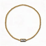 An April Birthstone White Cubic Zirconia Baguette Bracelet by Skōsh Jewelry, featuring a strand of polished gold beads on a stretch cord with a rectangular clear baguette stone at the center, displayed against a white background.