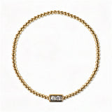 An April Birthstone White Cubic Zirconia Baguette Bracelet by Skōsh Jewelry, featuring a strand of polished gold beads on a stretch cord with a rectangular clear baguette stone at the center, displayed against a white background.
