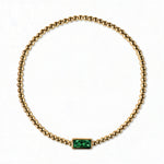 A May Birthstone Emerald Cubic Zirconia Baguette Bracelet by Skōsh Jewelry, featuring a strand of polished gold beads on a stretch cord with a rectangular green baguette stone at the center, displayed against a white background.