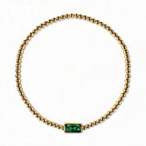 A May Birthstone Emerald Cubic Zirconia Baguette Bracelet by Skōsh Jewelry, featuring a strand of polished gold beads on a stretch cord with a rectangular green baguette stone at the center, displayed against a white background.