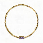 A June Birthstone Lavender Cubic Zirconia Baguette Bracelet by Skōsh Jewelry, featuring a strand of polished gold beads on a stretch cord with a rectangular light purple baguette stone at the center, displayed against a white background.