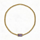 A June Birthstone Lavender Cubic Zirconia Baguette Bracelet by Skōsh Jewelry, featuring a strand of polished gold beads on a stretch cord with a rectangular light purple baguette stone at the center, displayed against a white background.