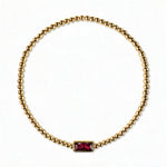 A July Birthstone Ruby Cubic Zirconia Baguette Bracelet by Skōsh Jewelry, featuring a strand of polished gold beads on a stretch cord with a rectangular red baguette stone at the center, displayed against a white background.