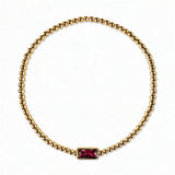A July Birthstone Ruby Cubic Zirconia Baguette Bracelet by Skōsh Jewelry, featuring a strand of polished gold beads on a stretch cord with a rectangular red baguette stone at the center, displayed against a white background.