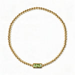 An August Birthstone Peridot Cubic Zirconia Baguette Bracelet by Skōsh Jewelry, featuring a strand of polished gold beads on a stretch cord with a rectangular light green baguette stone at the center, displayed against a white background.