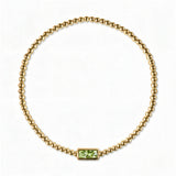 An August Birthstone Peridot Cubic Zirconia Baguette Bracelet by Skōsh Jewelry, featuring a strand of polished gold beads on a stretch cord with a rectangular light green baguette stone at the center, displayed against a white background.