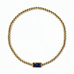 A September Birthstone Sapphire Cubic Zirconia Baguette Bracelet by Skōsh Jewelry, featuring a strand of polished gold beads on a stretch cord with a rectangular deep blue baguette stone at the center, displayed against a white background.