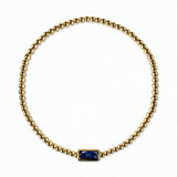 A September Birthstone Sapphire Cubic Zirconia Baguette Bracelet by Skōsh Jewelry, featuring a strand of polished gold beads on a stretch cord with a rectangular deep blue baguette stone at the center, displayed against a white background.