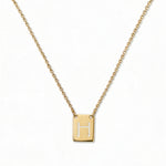 An Engraved Initial Necklace Letter H by Skōsh Jewelry, featuring a small rectangular tag engraved with the letter H on a fine gold chain, displayed against a white background.