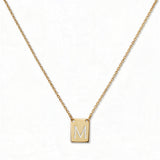 An Engraved Initial Necklace Letter M by Skōsh Jewelry, featuring a small rectangular tag engraved with the letter M on a fine gold chain, displayed against a white background.