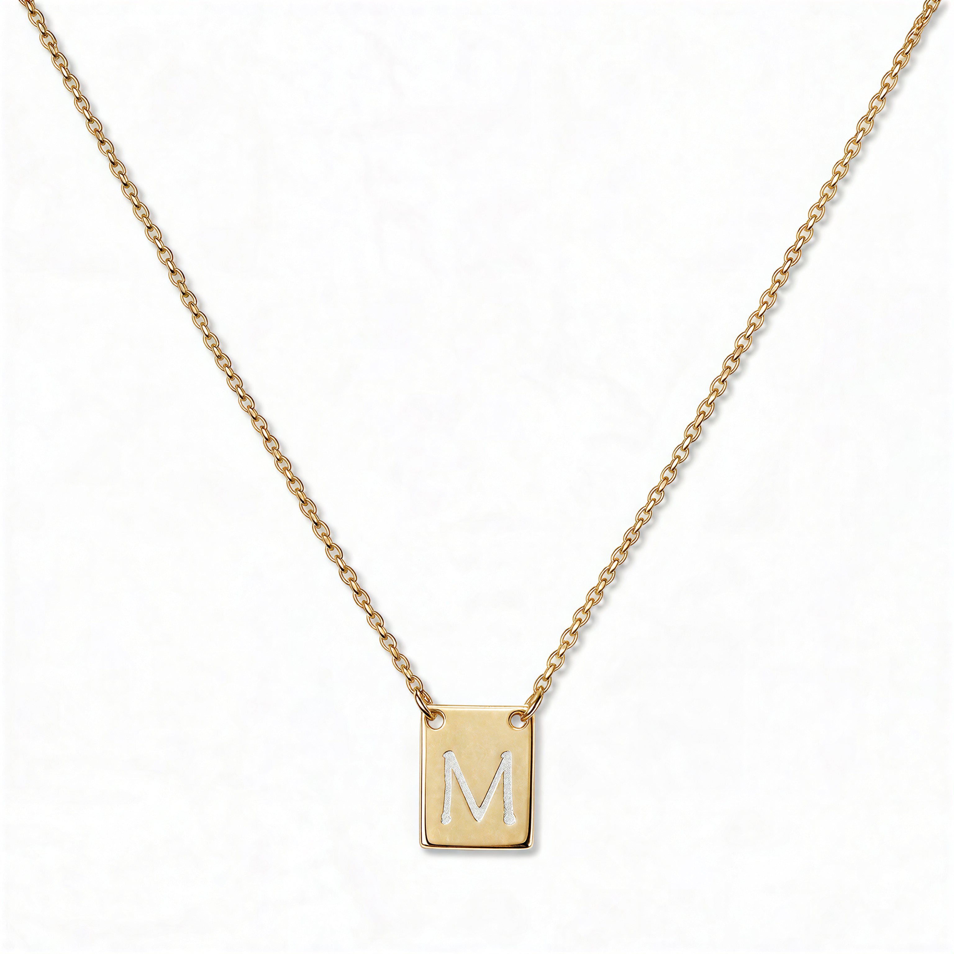 An Engraved Initial Necklace Letter M by Skōsh Jewelry, featuring a small rectangular tag engraved with the letter M on a fine gold chain, displayed against a white background.
