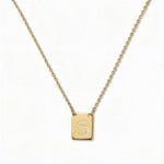 An Engraved Initial Necklace Letter S by Skōsh Jewelry, featuring a small rectangular tag engraved with the letter S on a fine gold chain, displayed against a white background.