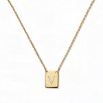 An Engraved Initial Necklace Letter V by Skōsh Jewelry, featuring a small rectangular tag engraved with the letter V on a fine gold chain, displayed against a white background.
